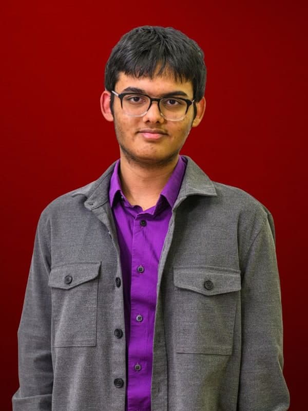 Abhimanyu Mishra in glasses and a grey overshirt over a purple shirt, against a red studio backdrop