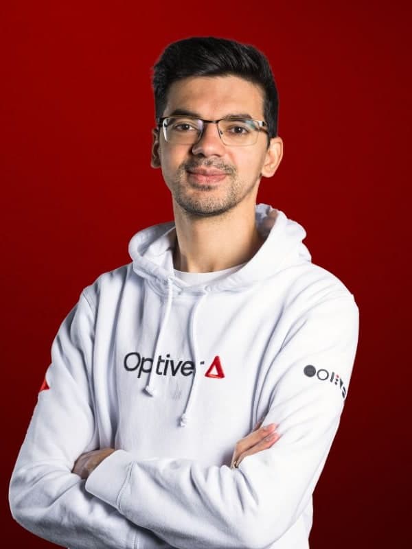 Anish Giri in glasses and a white hoodie, arms folded, against a red studio backdrop