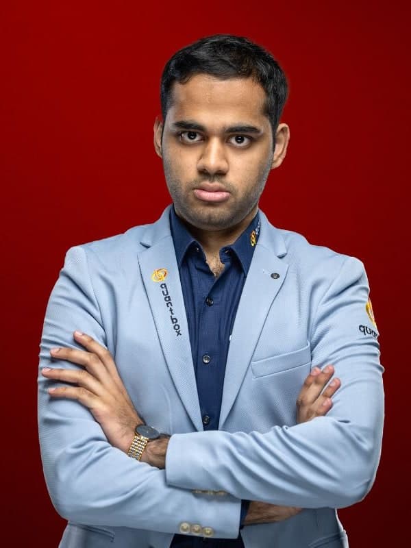 Arjun Erigaisi in a light blue blazer, arms folded, against a red studio backdrop