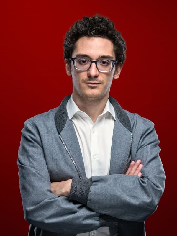 Fabiano Caruana in glasses and a grey jacket, arms folded, against a red studio backdrop