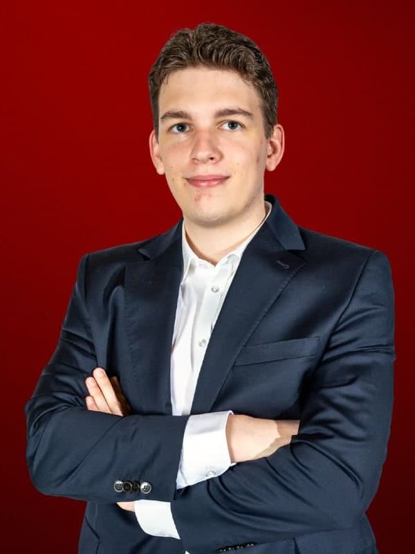 Jan-Krzysztof Duda in a navy suit, arms folded, against a red studio backdrop