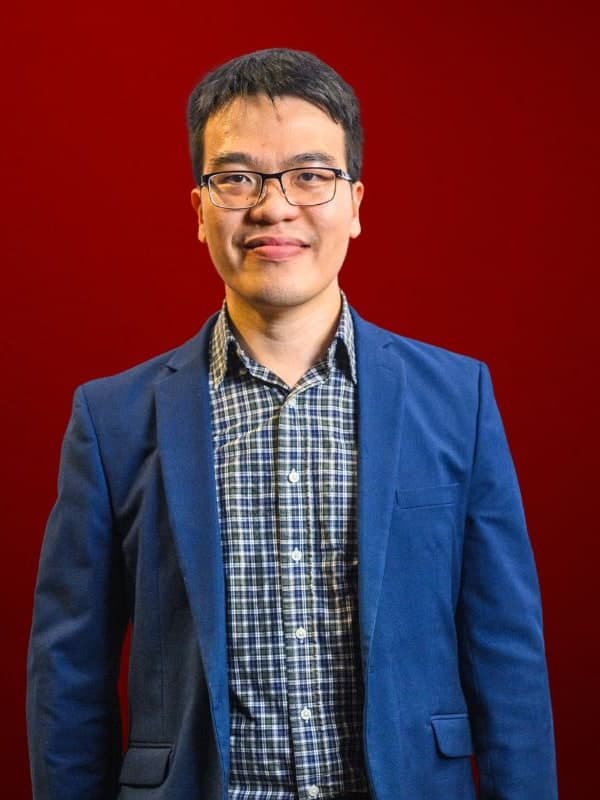 Liem Le in glasses, a blue blazer and a checked shirt, against a red studio backdrop