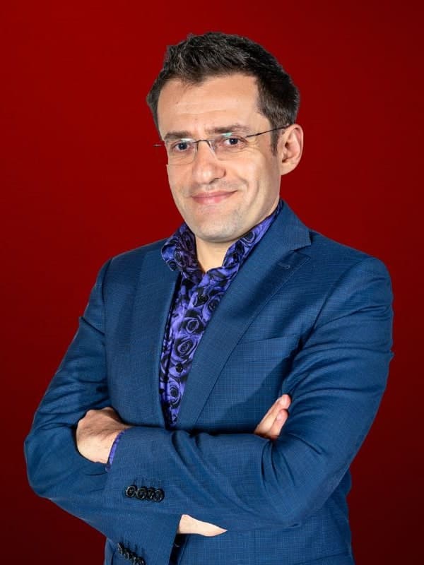 Levon Aronian in a blue suit and patterned shirt, arms folded, against a red studio backdrop