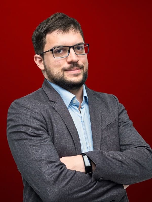 Maxime Vachier-Lagrave in glasses and a grey blazer, arms folded, against a red studio backdrop