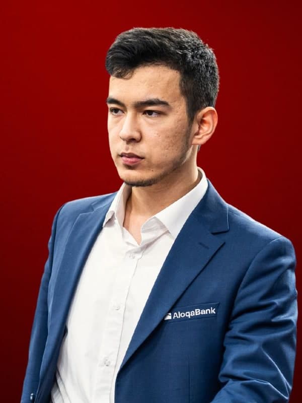 Nodirbek Abdusattorov in a blue blazer, looking to one side, against a red studio backdrop