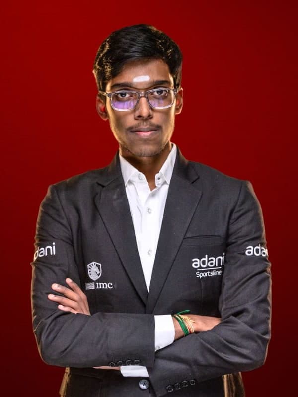 Praggnanandhaa R in glasses and a black team jacket, arms folded, against a red studio backdrop