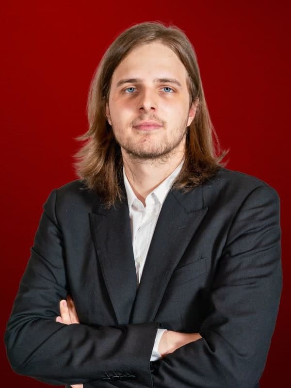 Richard Rapport, long-haired, in a black blazer, arms folded, against a red studio backdrop
