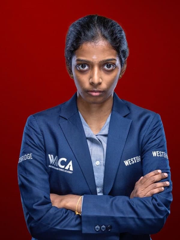 Vaishali Rameshbabu in a navy team blazer, arms folded, against a red studio backdrop