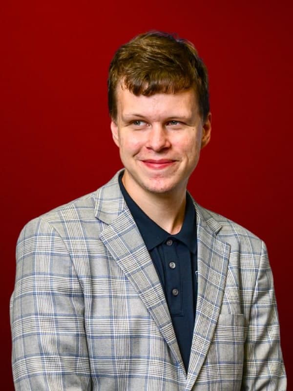Vladislav Artemiev, smiling, in a checked grey blazer, arms folded, against a red studio backdrop