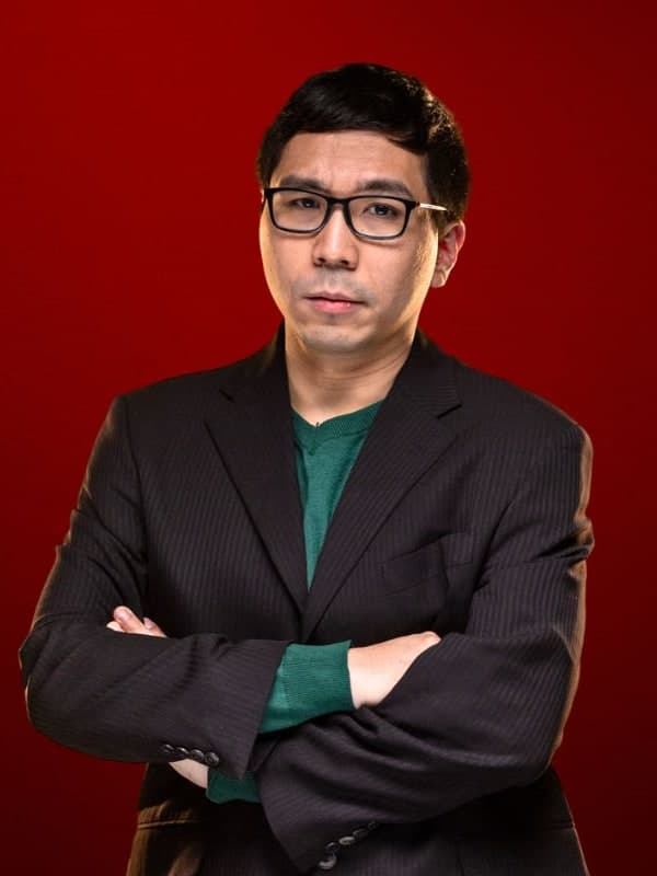 Wesley So in glasses and a dark blazer, arms folded, against a red studio backdrop